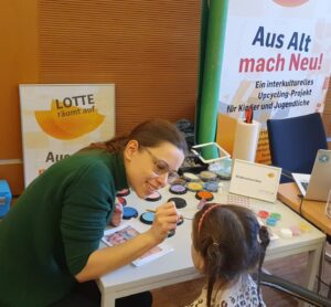 An Einem Kinderschmiktisch Wird Einem Mädchen Von Einer LOTTE-Mitarbeiterin Das Gesicht Geschminkt.
