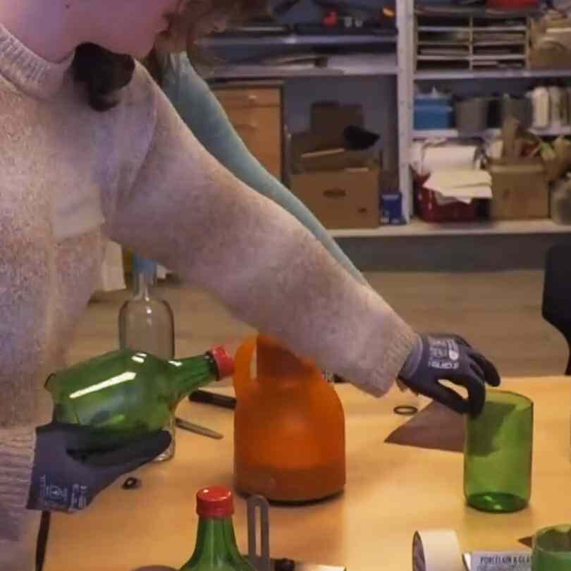 An einer Arbeitsplatte hält eine Jugendliche eine geschnittene Glasflasche. Ihre Hände sind mit Handschuhen geschützt.